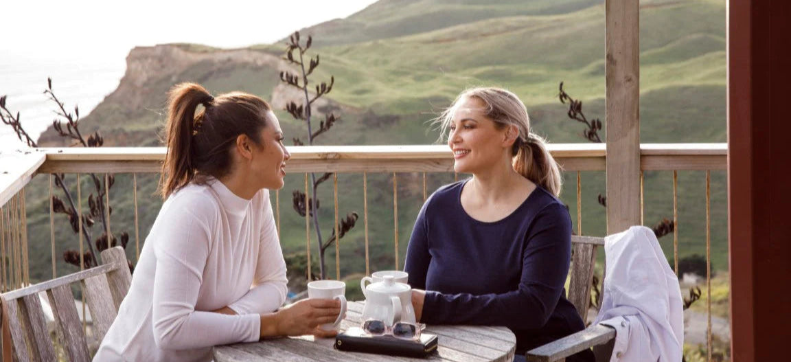 Two women wearing Merino Wool Jumpers sitting at a table on a balcony with a scenic mountain view.