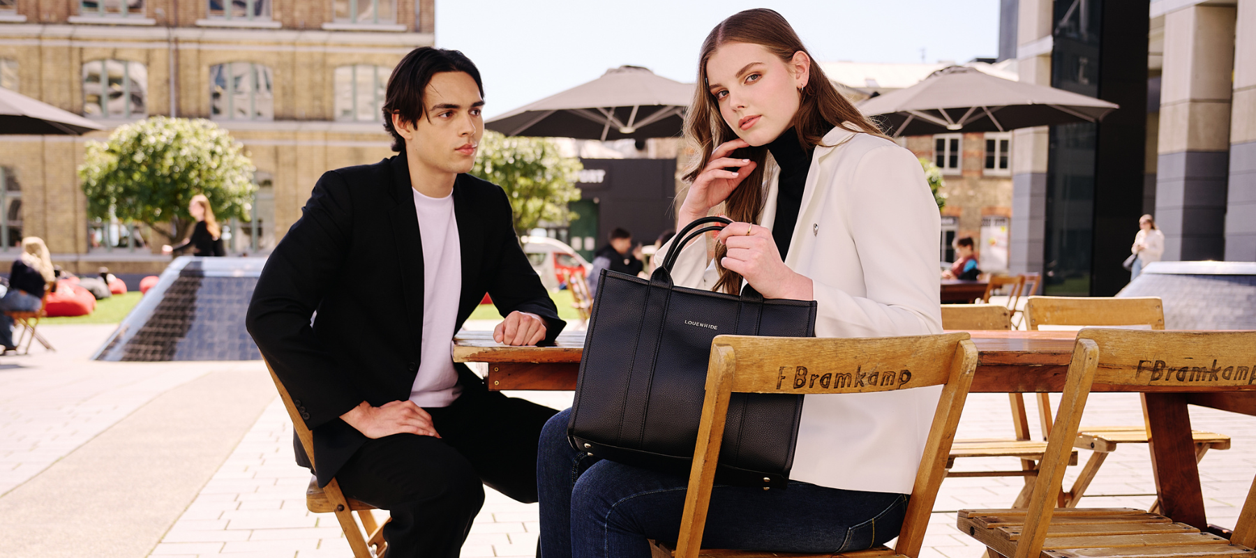A stylish woman and man wearing Ebony Boutique Clothes. She has a black high neck merino wool jumper on with an Ivory Gordon Smith Blazer and Denim Jeans . She is holding a Louenhide Manhattan Laptop Bag. 
