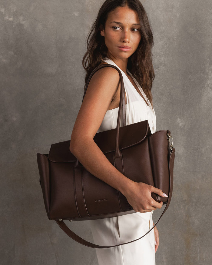 Woman carrying chocolate Leighton tote with matching Liv laptop sleeve styled for workday or business setting